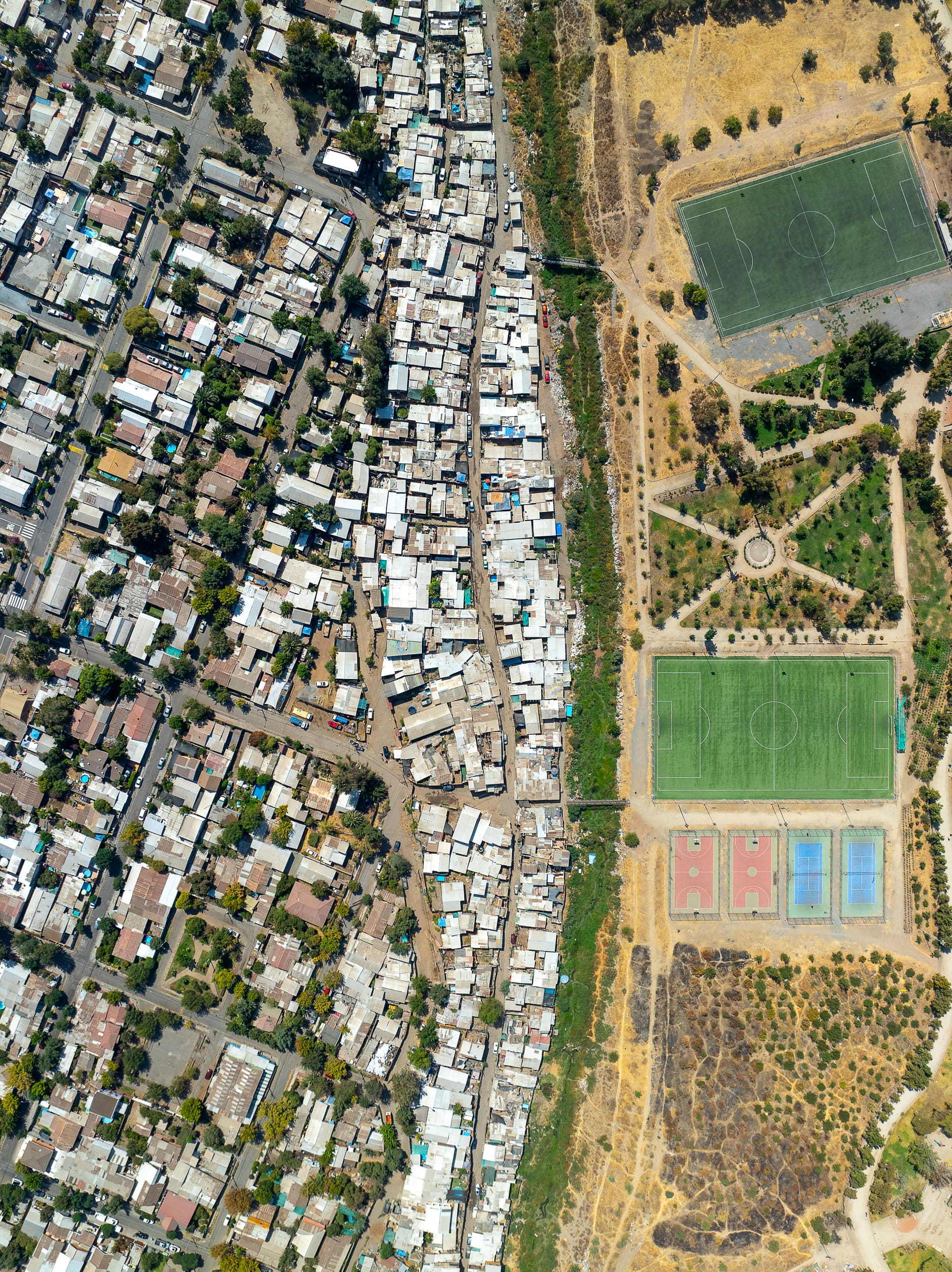 Santiago from directly overhead: dense self-built housing on one side of a channel, watered sports fields and planted parkland on the other
