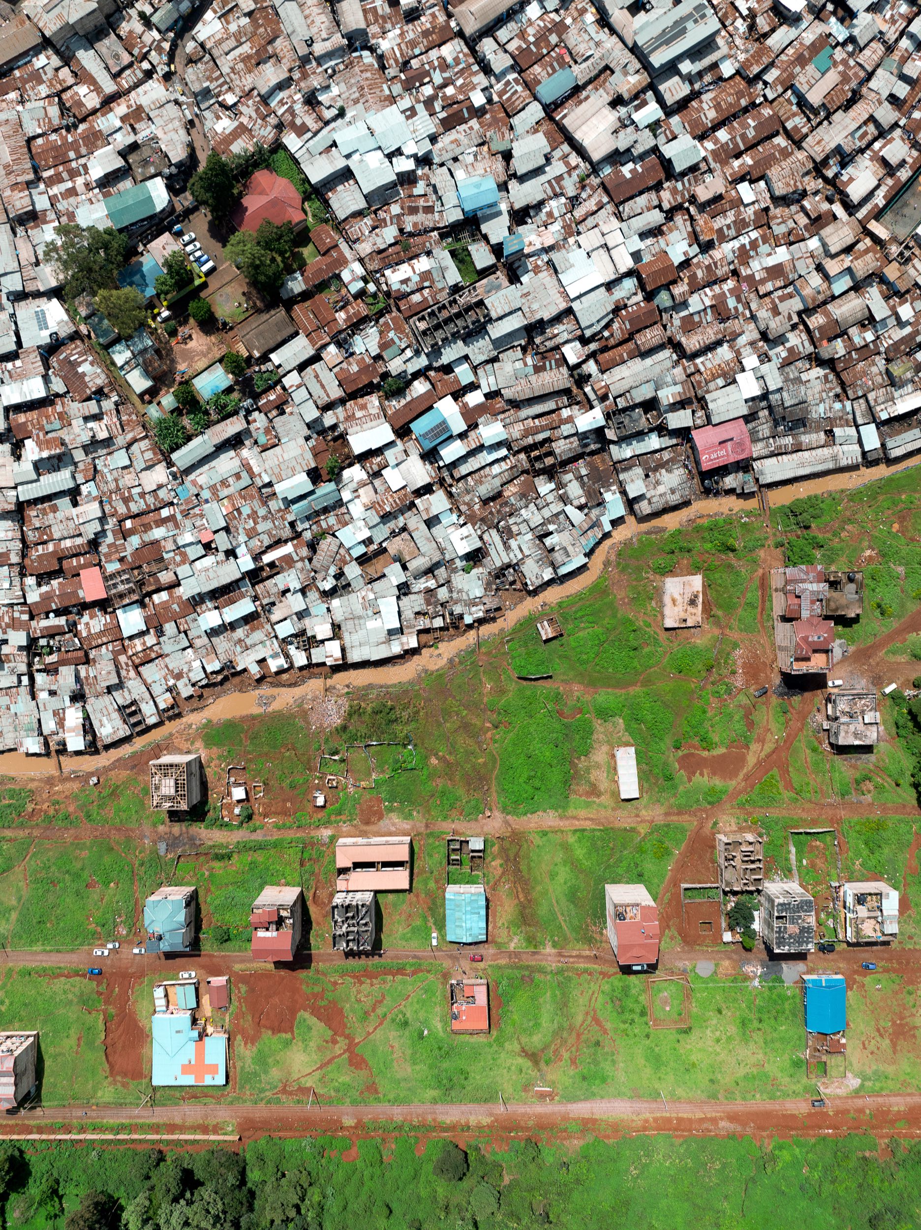 Nairobi from directly overhead: corrugated iron roofs packed edge to edge across the top of the frame, and below them open red earth, scattered half-built plots, trees, and a clinic with a red cross painted on its roof